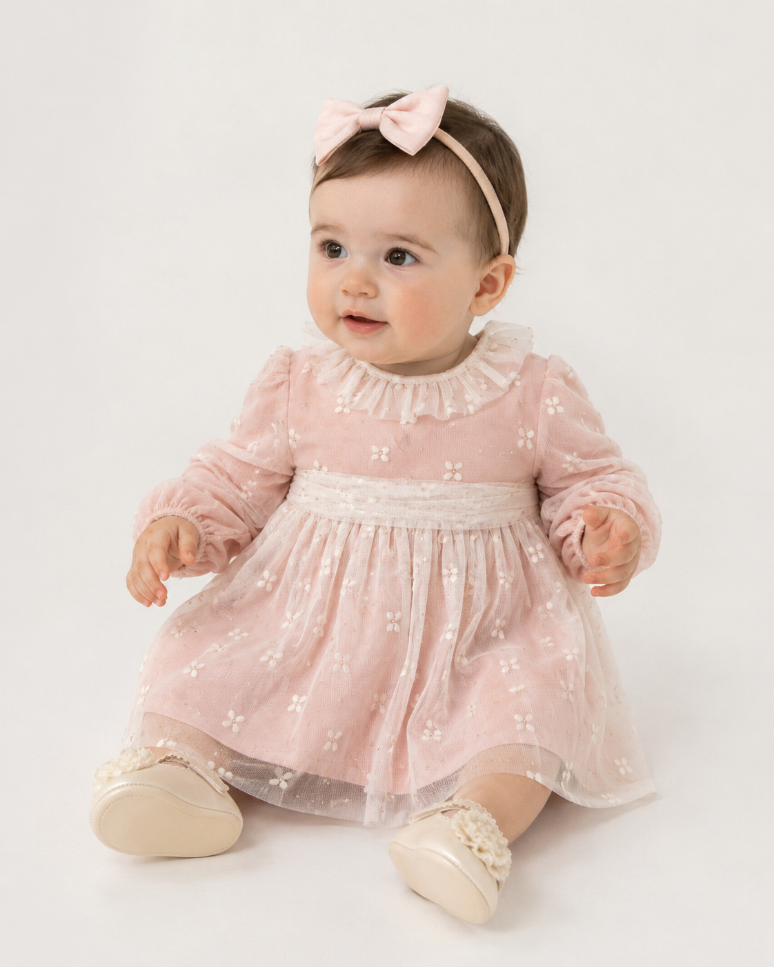 A baby girl wearing a light pink long-sleeve dress with cream butterfly embroidery, a ruffled collar, and a matching pink bow headband, sitting against a plain white studio background.