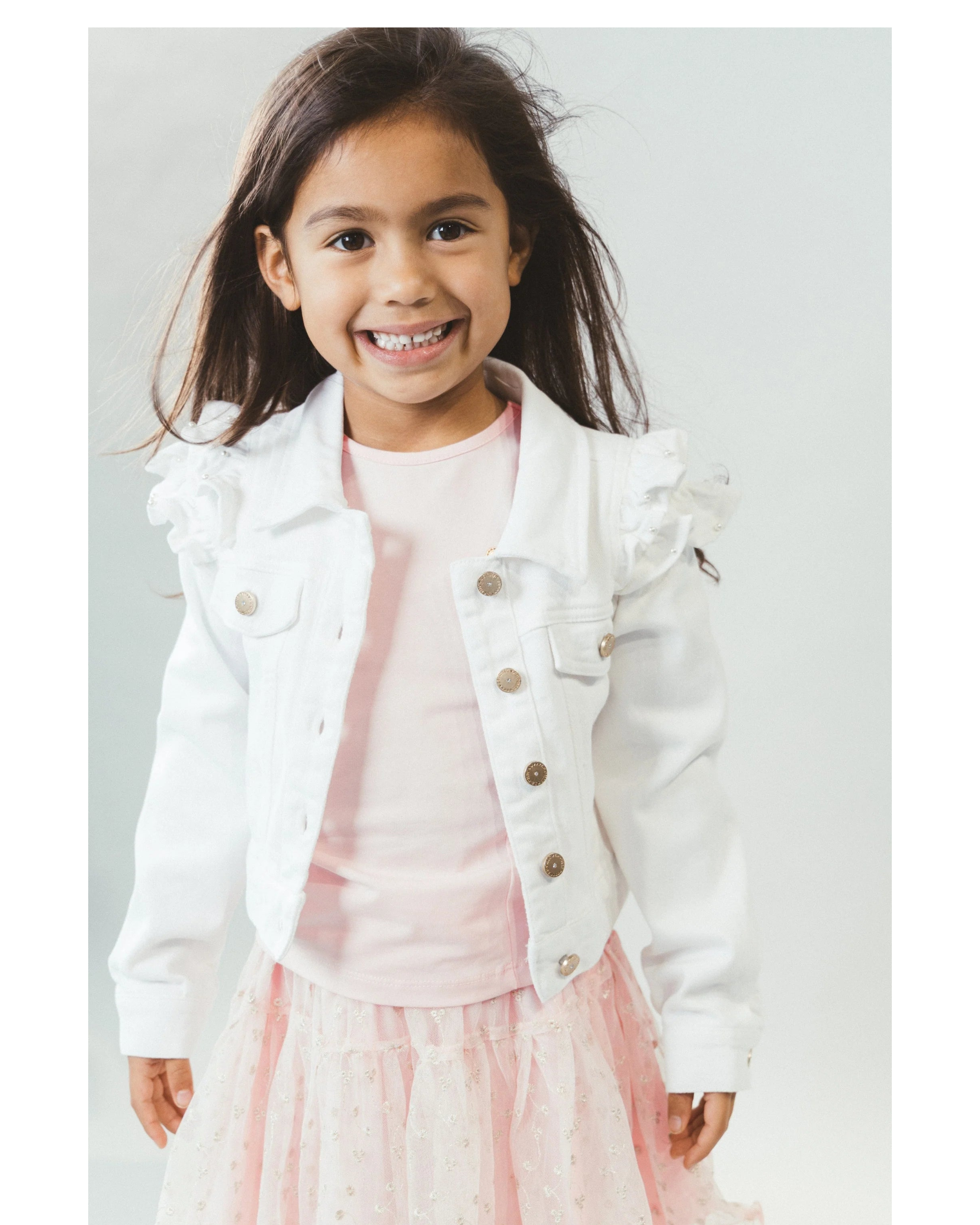 Young girl wearing a white jean  jacket over a pink dress on a light gray background