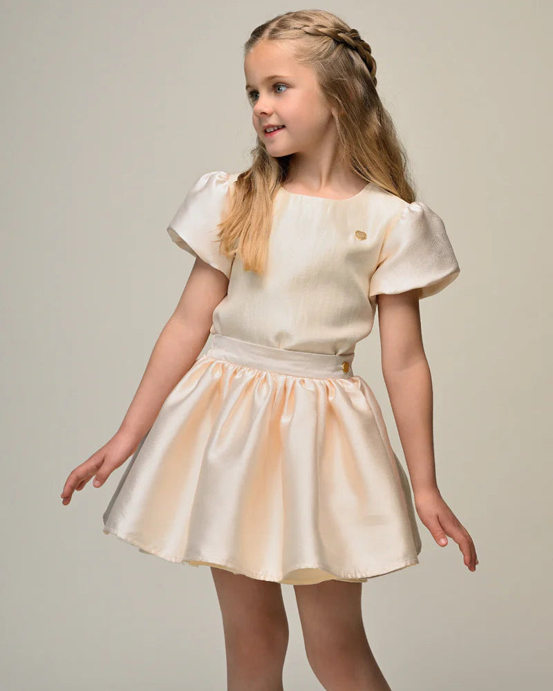 Young girl wearing a beige skirt and top  with puffed sleeves against a plain background