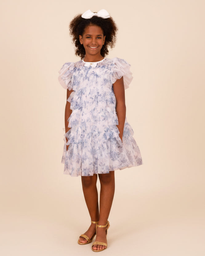 Young girl wearing a floral dress standing against a beige wall.