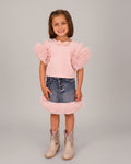 Young girl wearing a pink dress with ruffled sleeves and denim skirt, standing on a wooden floor.
