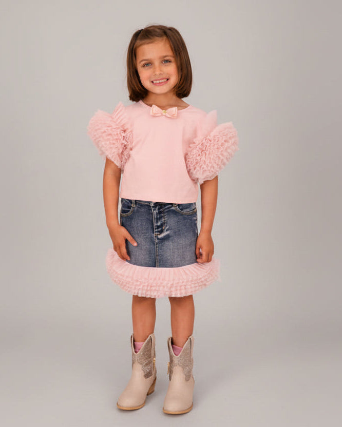 Young girl wearing a pink dress with ruffled sleeves and denim skirt, standing on a wooden floor.