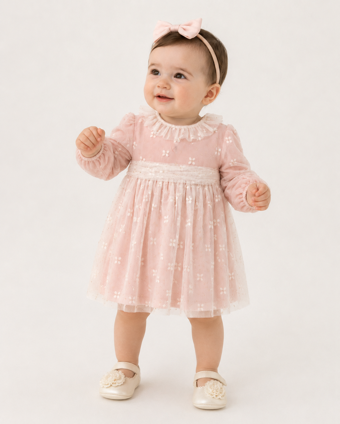 A baby girl wearing a light pink long-sleeve tulle dress with a ruffled collar and white embroidered floral details, paired with a matching pink bow headband and cream-colored shoes, standing against a plain white studio background.